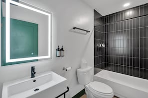 A modern bathroom with a white sink, toilet, and bathtub. There is a mirror with a green door, and black tiles on the wall.