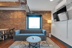 A room with a brick wall, a blue-framed window, a grey sofa, a small round table, and a TV mounted on the wall.