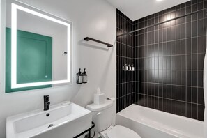 Uma casa de banho moderna com uma pia, sanita e banheira brancas, um espelho com porta verde e paredes revestidas a mosaico preto.