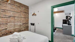 Ein Hotelzimmer mit Backsteinwand, einem Bett mit weißer Bettwäsche, einem Kleiderständer, einem aufgehängten Mantel und Blick in den Küchenbereich.
