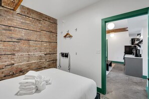 Quarto de hotel com parede de tijolos, cama com lençóis brancos, um cabideiro, um casaco pendurado e vista para a área da cozinha.