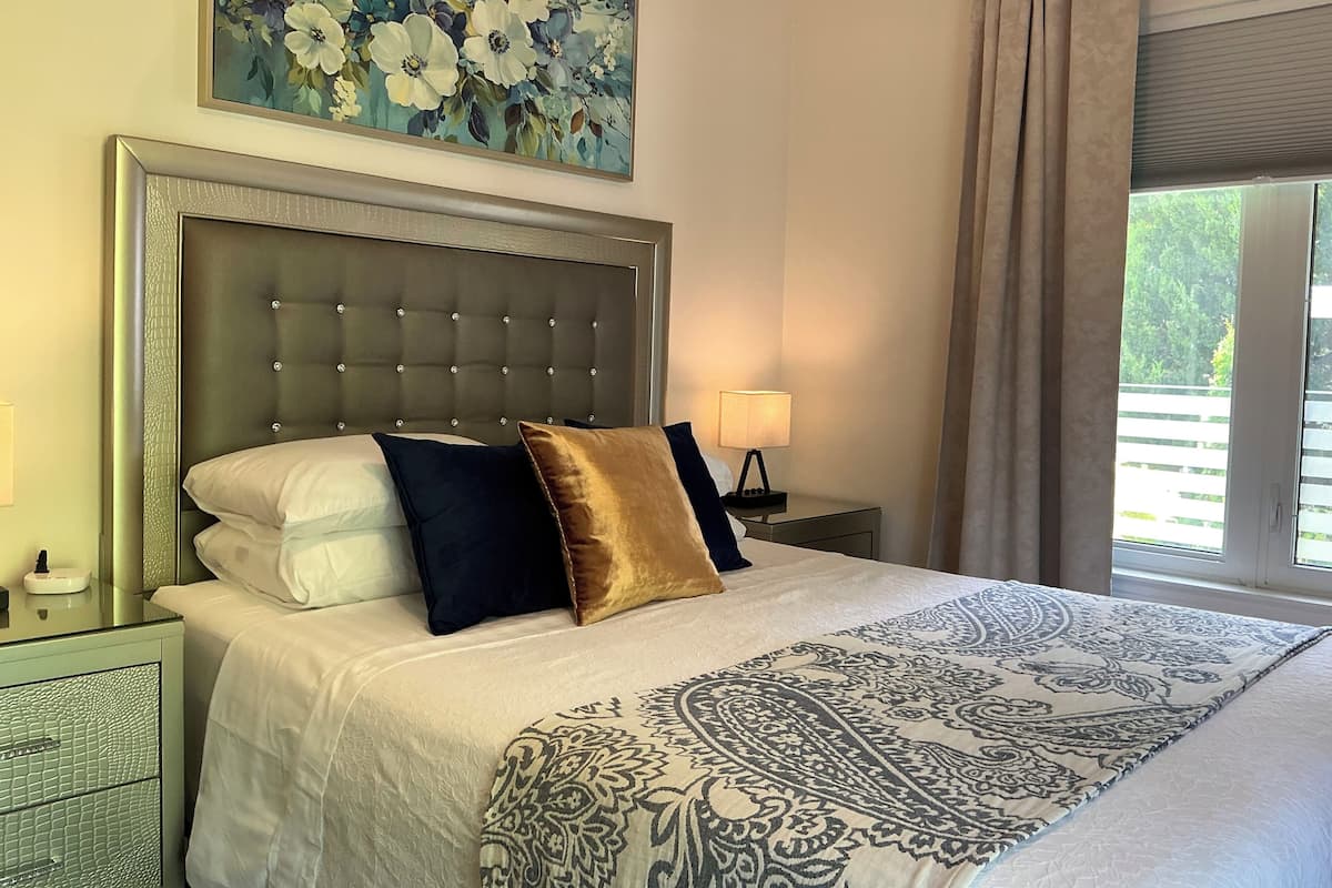 A hotel room with a large bed, a headboard, a nightstand, a lamp, a window with curtains, and a floral painting on the wall.