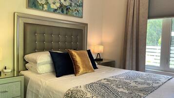 Ein Hotelzimmer mit einem großen Bett, einem Kopfteil, einem Nachttisch, einer Lampe, einem Fenster mit Vorhängen und einem floralen Gemälde an der Wand.