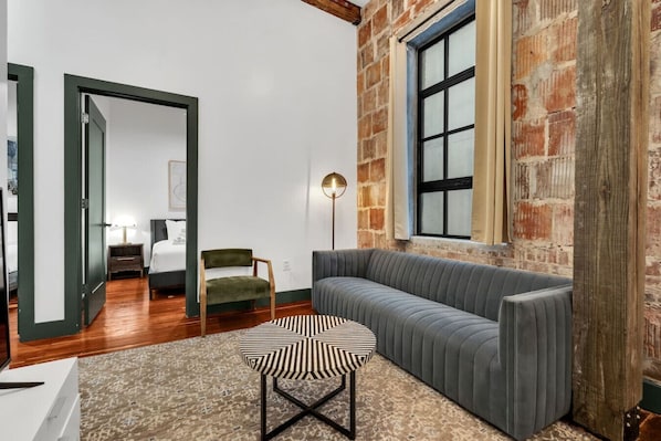 Uma sala de estar moderna com um sofá cinzento, um banquinho estampado, uma poltrona verde, um abajur e uma parede de tijolos.