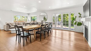 A spacious living area with a wooden dining table, black chairs, a round mirror, and a large window with a view of greenery. - Peaceful Single-Level Modern Luxury with Private Yard and AC in La Jolla (San Diego)