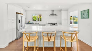A modern kitchen with white cabinetry, a large island with bar stools, stainless steel appliances, and a view of greenery outside. - Peaceful Single-Level Modern Luxury with Private Yard and AC in La Jolla (San Diego)