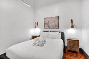 A neatly made bed with a white comforter and pillows, two bedside tables with lamps, a wooden floor, and a painting on the wall.