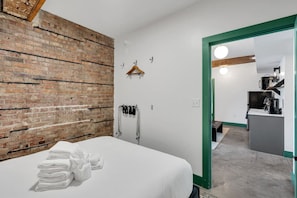 Quarto de hotel com parede de tijolos, cama com lençóis brancos, um cabideiro, um casaco pendurado e vista para a área da cozinha.