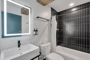 Um banheiro moderno com pia, vaso sanitário e banheira brancos, um espelho grande e paredes revestidas de azulejos pretos.