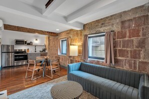 A modern kitchen with stainless steel appliances, a dining area with a wooden table and chairs, a blue sofa, and a brick wall.