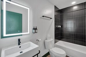 Un bany modern amb un lavabo, vàter i banyera de color blanc. Hi ha un mirall amb una porta verda, i rajoles negres a la paret.