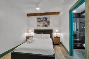 Quarto com cama, duas mesas de cabeceira com luminárias, um vigamento de madeira no teto, um quadro na parede e um banheiro visível através de uma porta aberta.