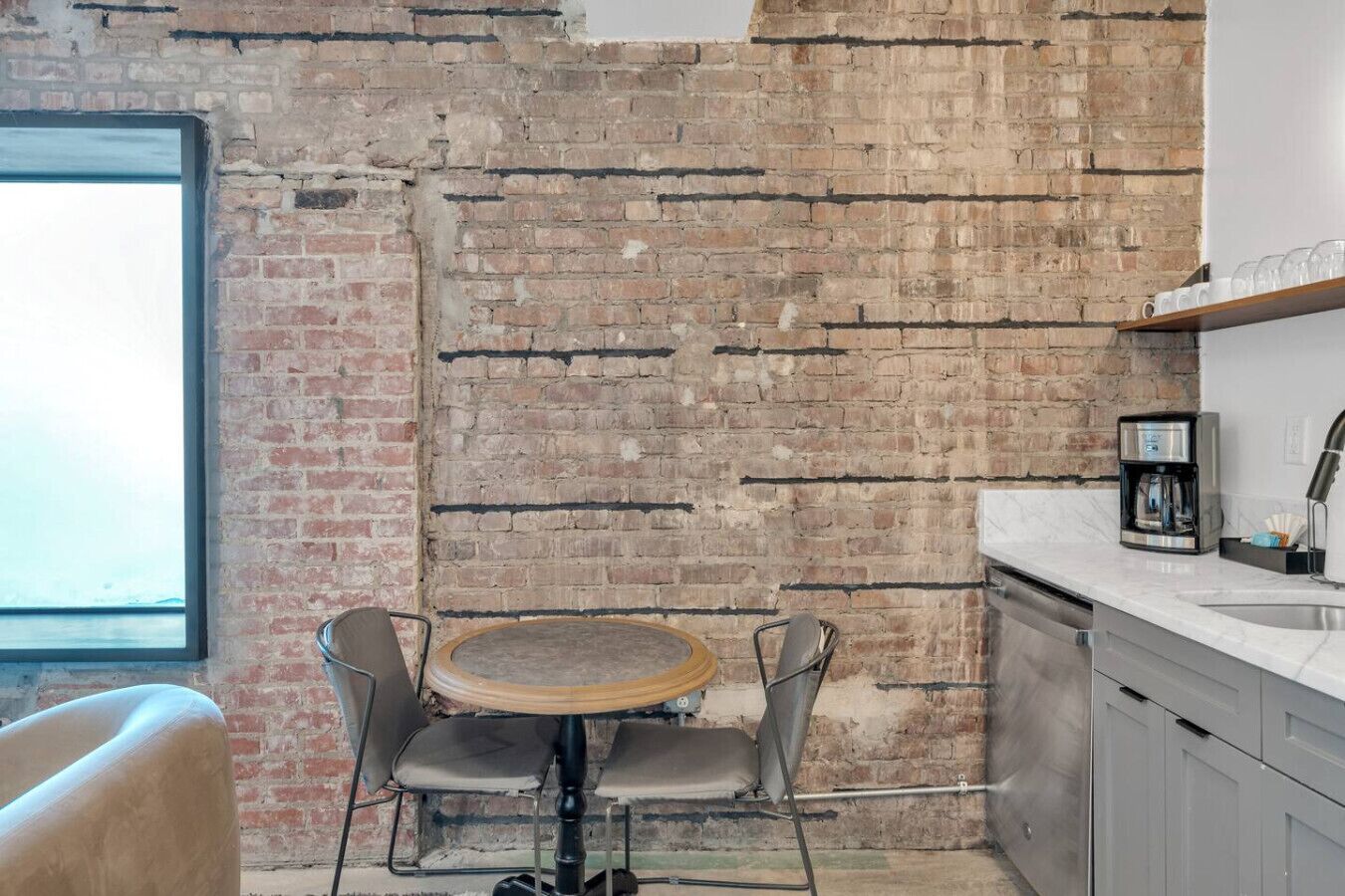 Una cocina con pared de ladrillo, una mesa redonda con sillas, una cafetera y una ventana.