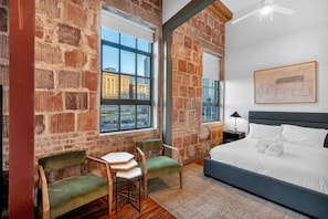 A bedroom with a brick wall, a large window, a bed, a desk, and two chairs.