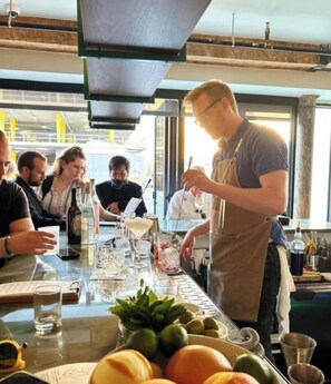 Uma área de bar com um bartender preparando bebidas, um menu e uma mesa com copos e garrafas.
