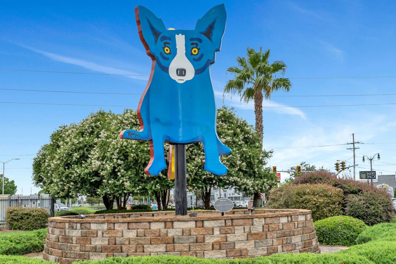 Una grande scultura di un cane blu su un piedistallo, in un’area verdeggiante con alberi e un muro di mattoni.