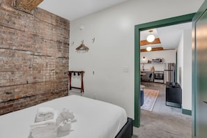 A bedroom with a brick wall, a bed with white linens, a wooden hanger, and a view into a kitchen area.