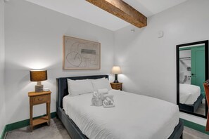 Uma cama bem arrumada com lençóis brancos, duas luminárias de cabeceira, uma mesinha de cabeceira de madeira, uma obra de arte abstrata emoldurada e um espelho grande refletindo uma porta verde.