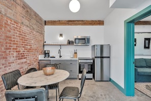 A compact kitchen with a brick wall, a dining area with a table and chairs, a microwave, a refrigerator, and a coffee maker.