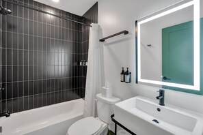 Uma casa de banho moderna com banheira, sanita e lavatório, com azulejos escuros, porta verde e espelho com moldura iluminada.