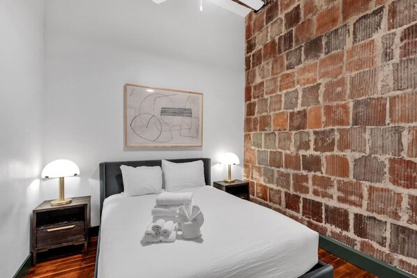A bedroom with a brick wall, a bed with white linens, two bedside tables with lamps, and a framed abstract artwork on the wall.