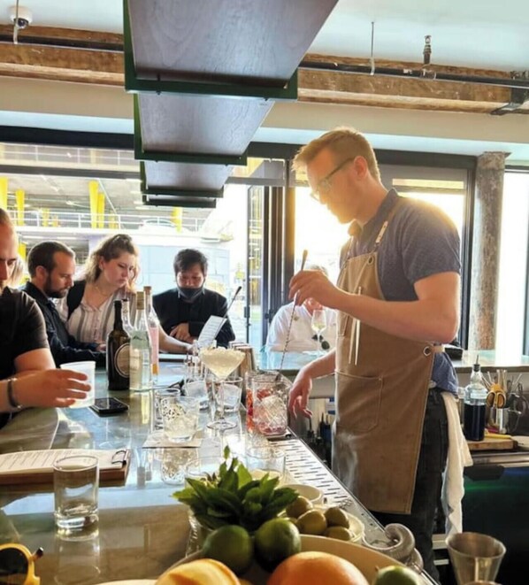 A bar area with a bartender serving drinks, a table with glasses and bottles, and a window with a view of the outside.