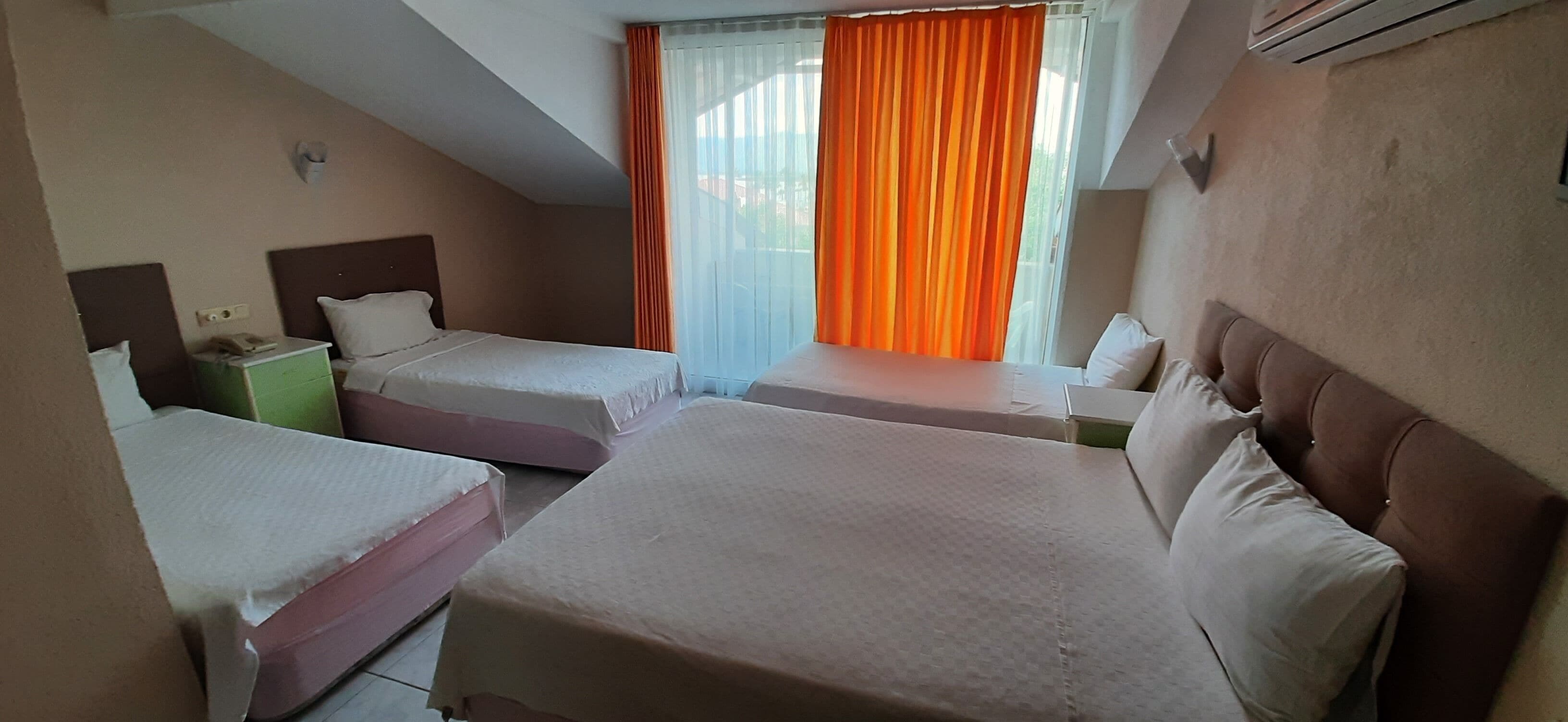 A hotel room with two beds, a small table, a nightstand, and a window with orange curtains.