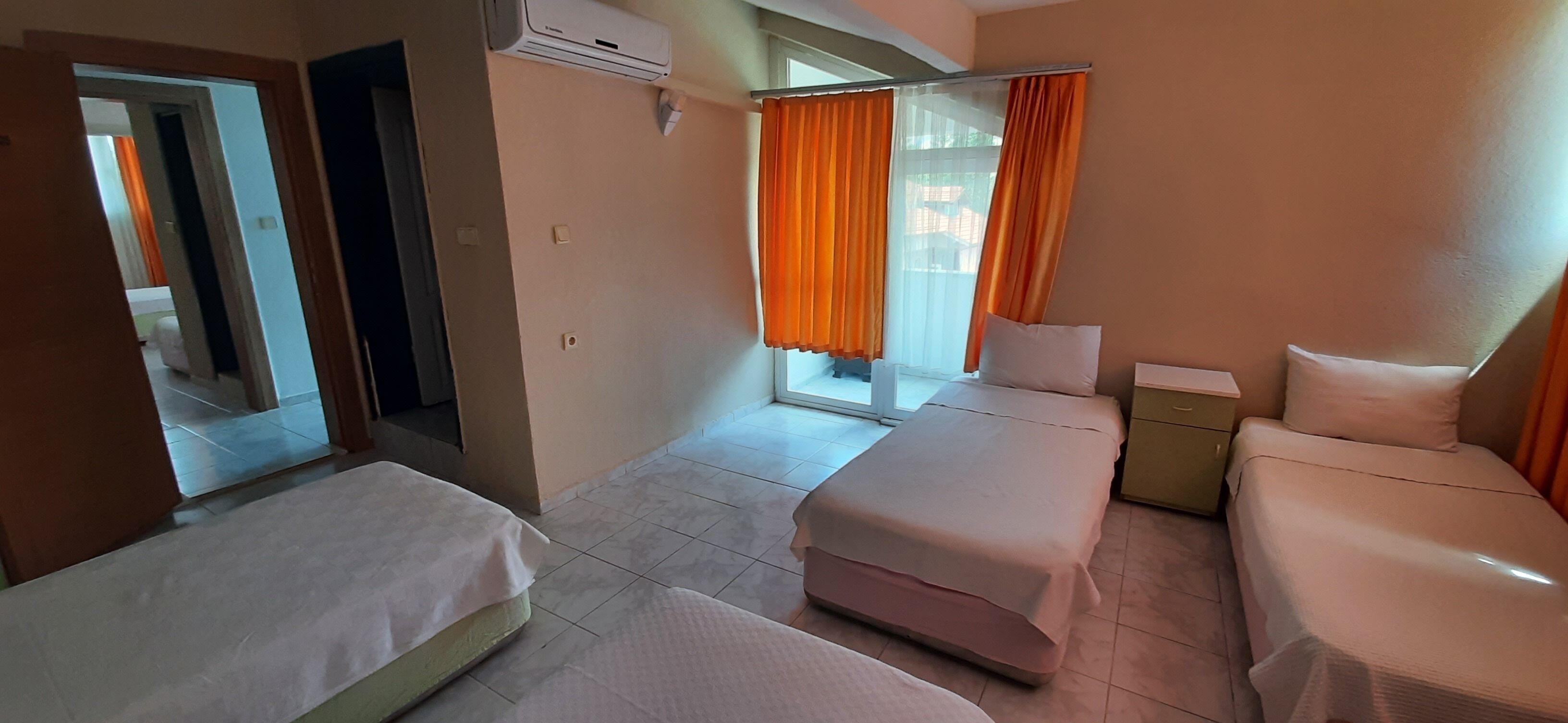 A hotel room with two beds, a small table, an air conditioning unit, and a window with orange curtains.