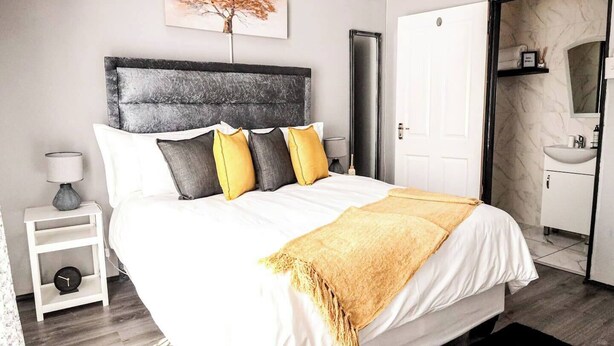 A modern bedroom with a large bed, a headboard, bedside tables, a nightstand, a clock, a painting, a door, and a bathroom with a sink and mirror. - Elite Luxury Garden Escape, Executive Cottage in Welkom with Wi-Fi & Parking (Welkom)