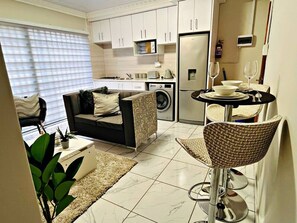 A compact living space with a sofa, dining table, and chairs, a washing machine, and a refrigerator.