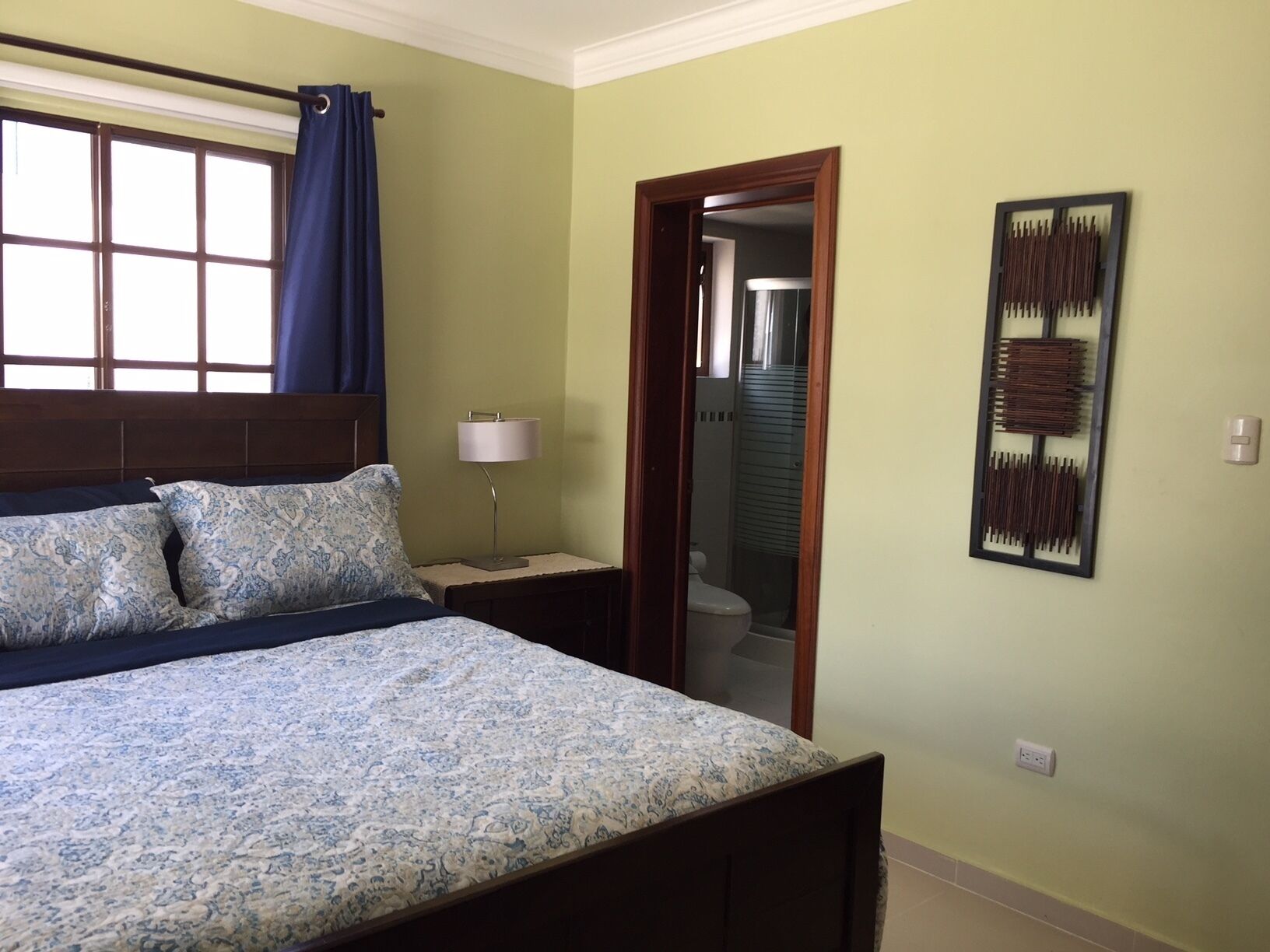 A bedroom with a bed, a nightstand, a lamp, a window with curtains, a door, and a wall-mounted artwork.