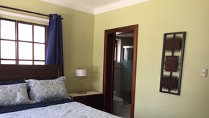 Une chambre à coucher comprenant un lit, une table de chevet, une lampe, une fenêtre avec des rideaux, une porte et une œuvre d’art accrochée au mur.