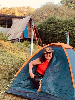 A person sitting inside a tent with a blue and orange exterior, surrounded by greenery and a wooden structure in the background. - Camping Monte Verde  (Monte Verde)