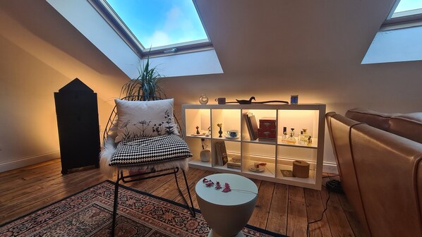 A room with a skylight, a chair with a patterned cushion, a small table, and a shelf with various items.