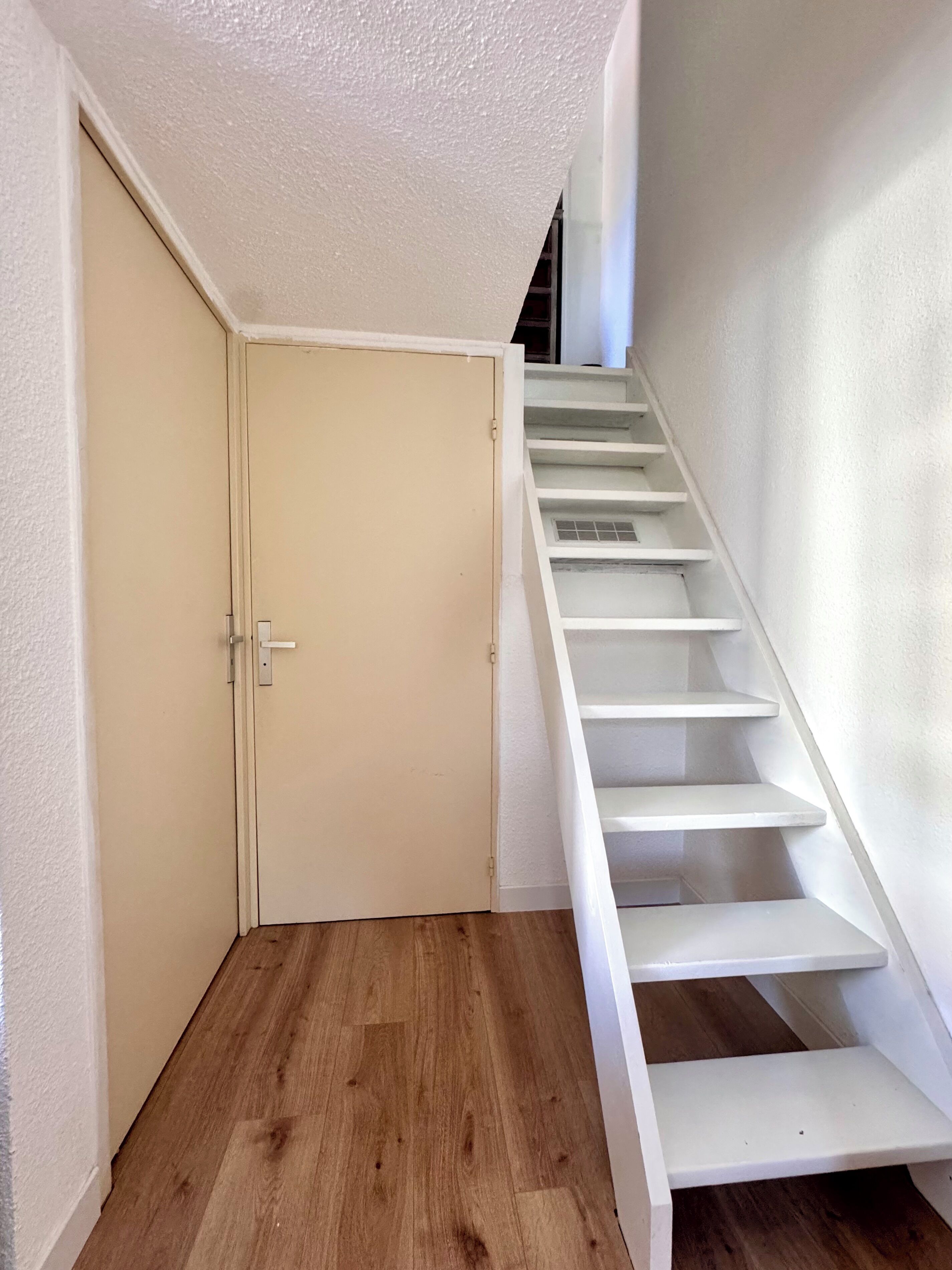A staircase with white steps leading to an upper floor, a closed door with a handle, and a wooden floor.