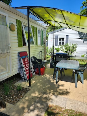 Ein überdachter Patio-Bereich mit Tisch und Stühlen, einem Grill und einem Wohnwagen mit grünen Fensterläden.
