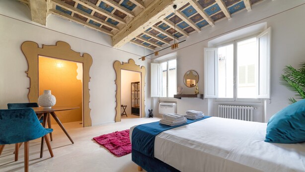 Quarto com cama, poltrona, radiador, janela e teto com vigas aparentes.