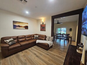 A living room with a brown leather sectional sofa, wooden flooring, a dining area with a table and chairs, and a large TV screen.