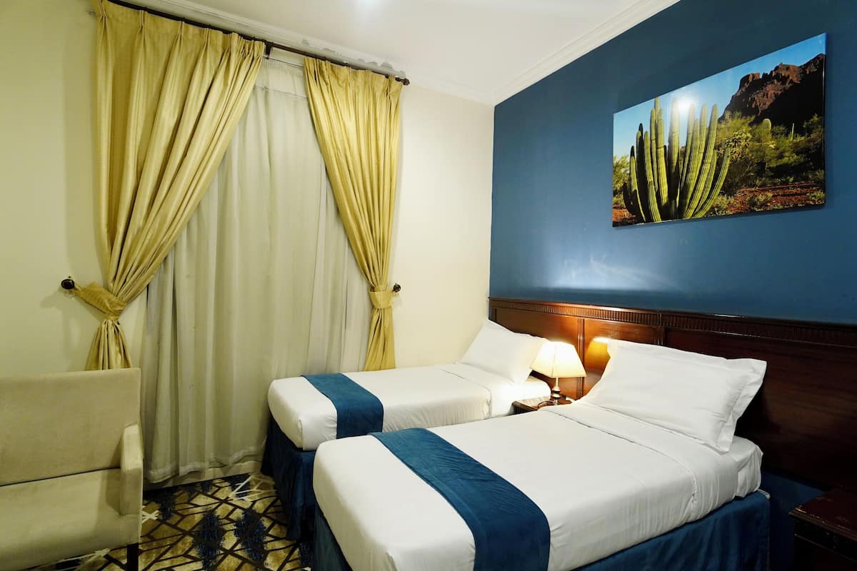 A hotel room with two beds, a chair, a nightstand with a lamp, and a large cactus painting on the wall.