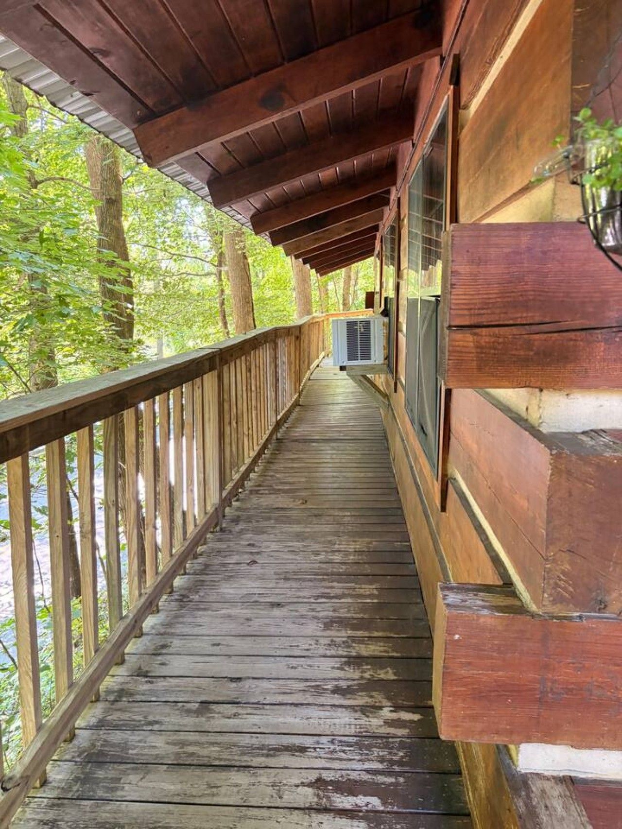 A wooden deck with a railing, leading to a building with large windows and a metal air conditioning unit.