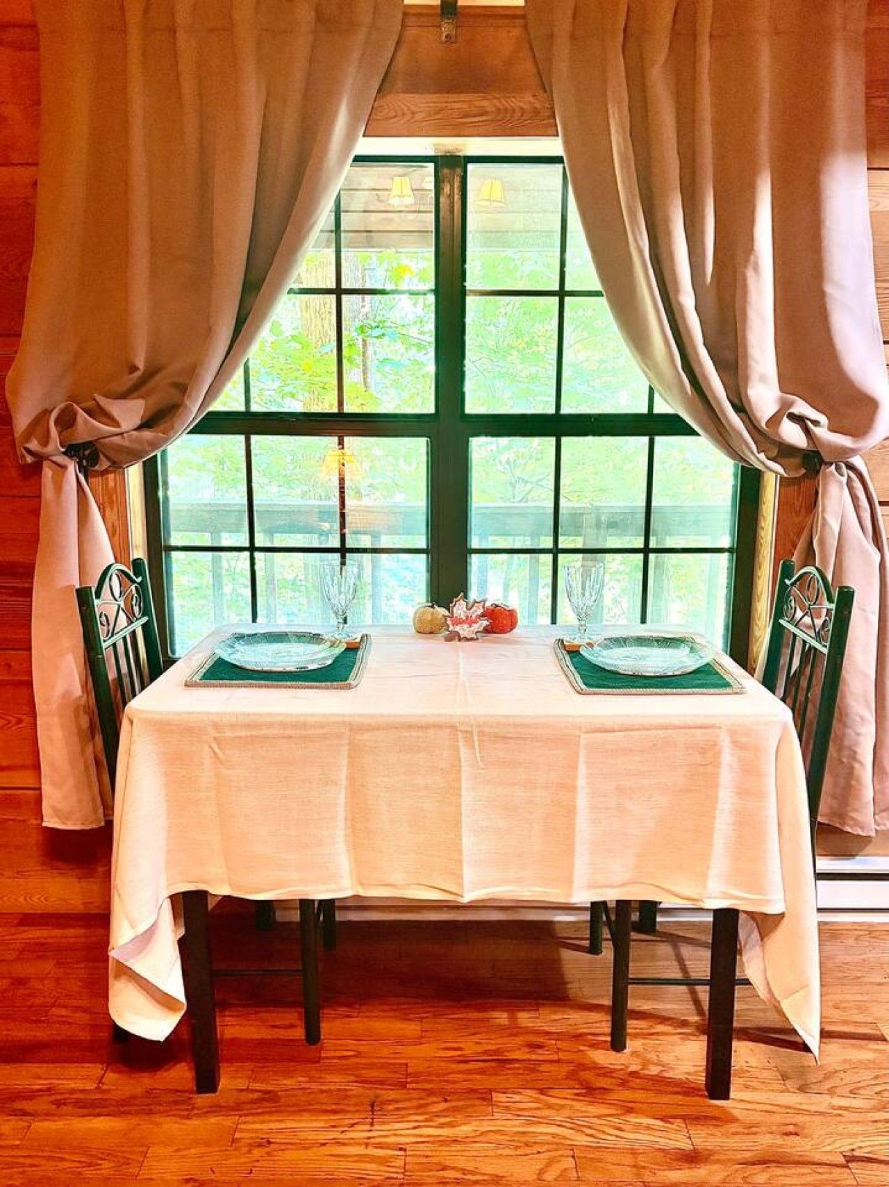 A dining table set for two with a white tablecloth, green placemats, and a view through a green-framed window.