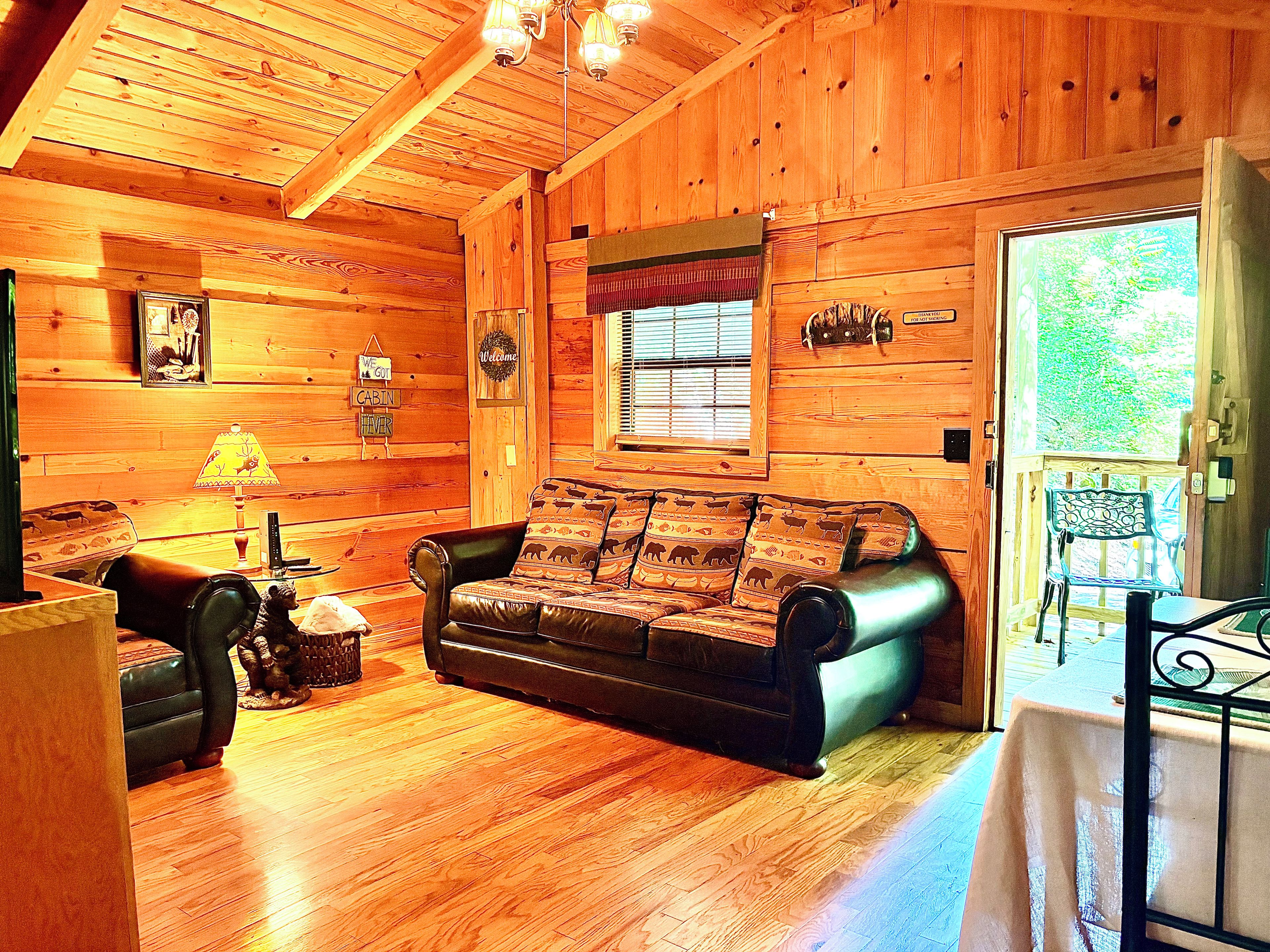 A cabin interior with a wooden floor, a leather sofa, a table with a lamp, and a window with blinds.