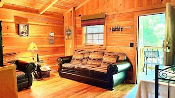 A cabin interior with a wooden floor, a leather sofa, a table with a lamp, and a window with blinds.