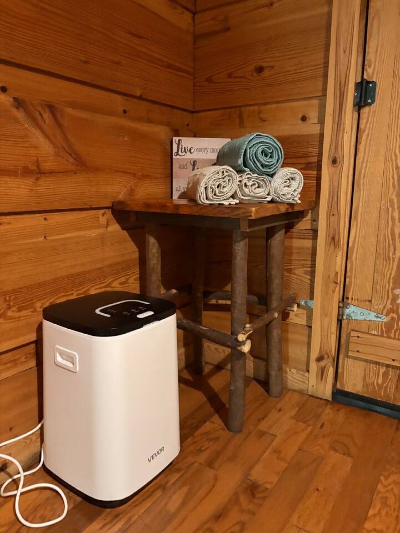 A wooden cabin interior with a white heater, a small table with rolled towels, and a sign with a motivational quote.