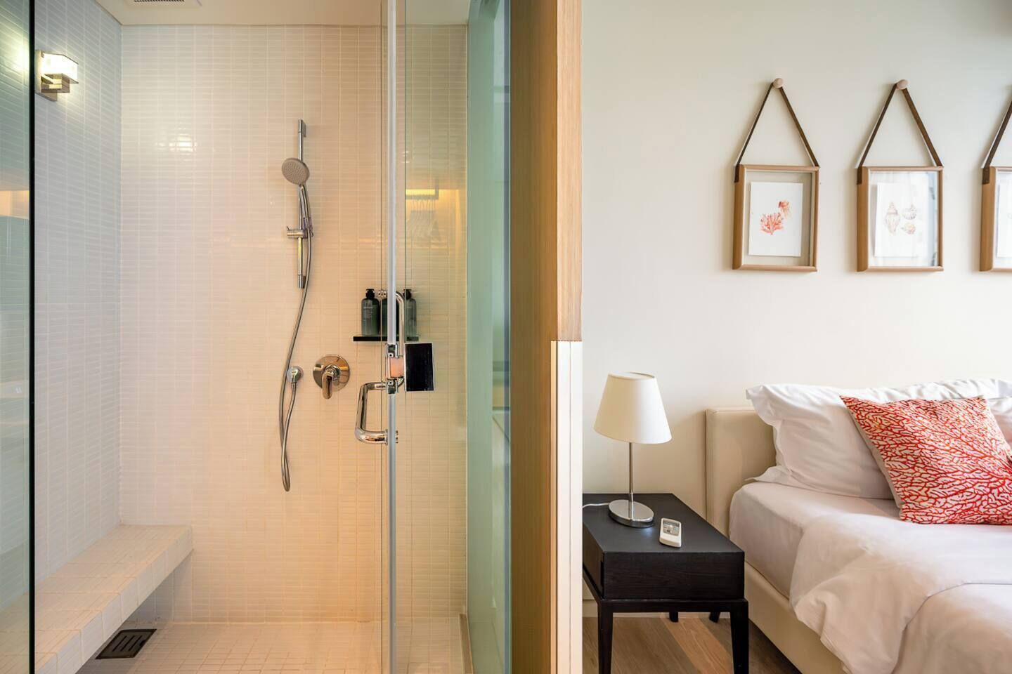 Un baño moderno con cabina de ducha de vidrio, una mesita de noche con lámpara y una cama con almohadas.