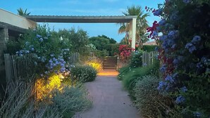 Un sendero pavimentado que conduce a una entrada con puerta, rodeado de abundante vegetación y plantas florecientes.