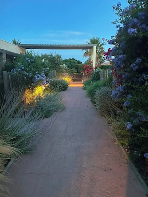 A paved pathway leading to a gated entrance, surrounded by lush greenery and flowering plants. - Apartment ROSSA - Standing, swimming pool & Cala Rossa beaches on foot (Lecci)