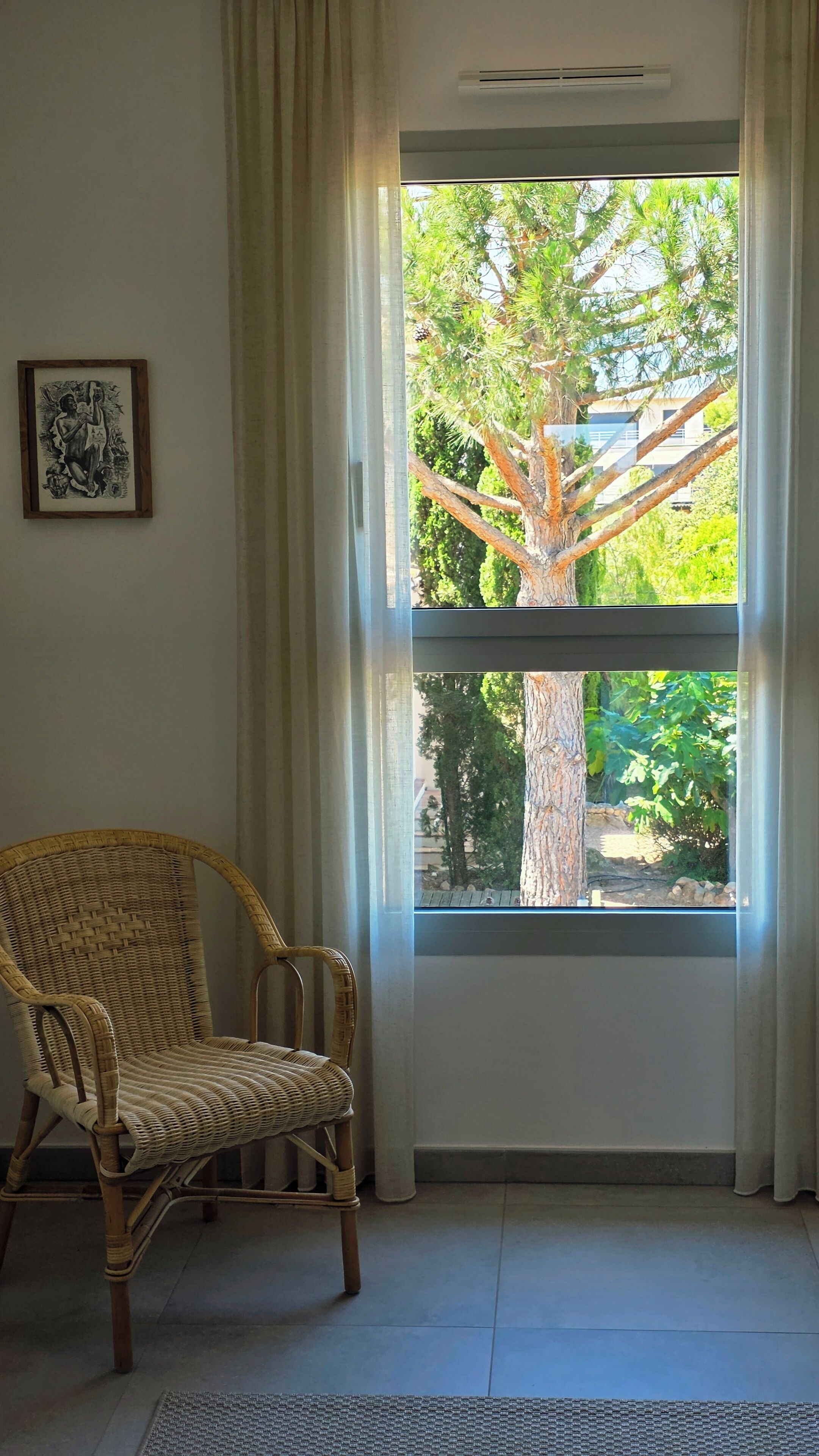 Ein Zimmer mit einem Korbstuhl, einem gerahmten Bild an der Wand und einem großen Fenster mit Blick auf Bäume und ein Gebäude.