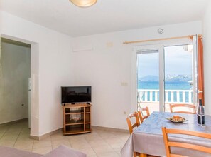 Quarto com televisão sobre suporte de madeira, mesa de jantar com cadeiras e varanda com vista para o mar.