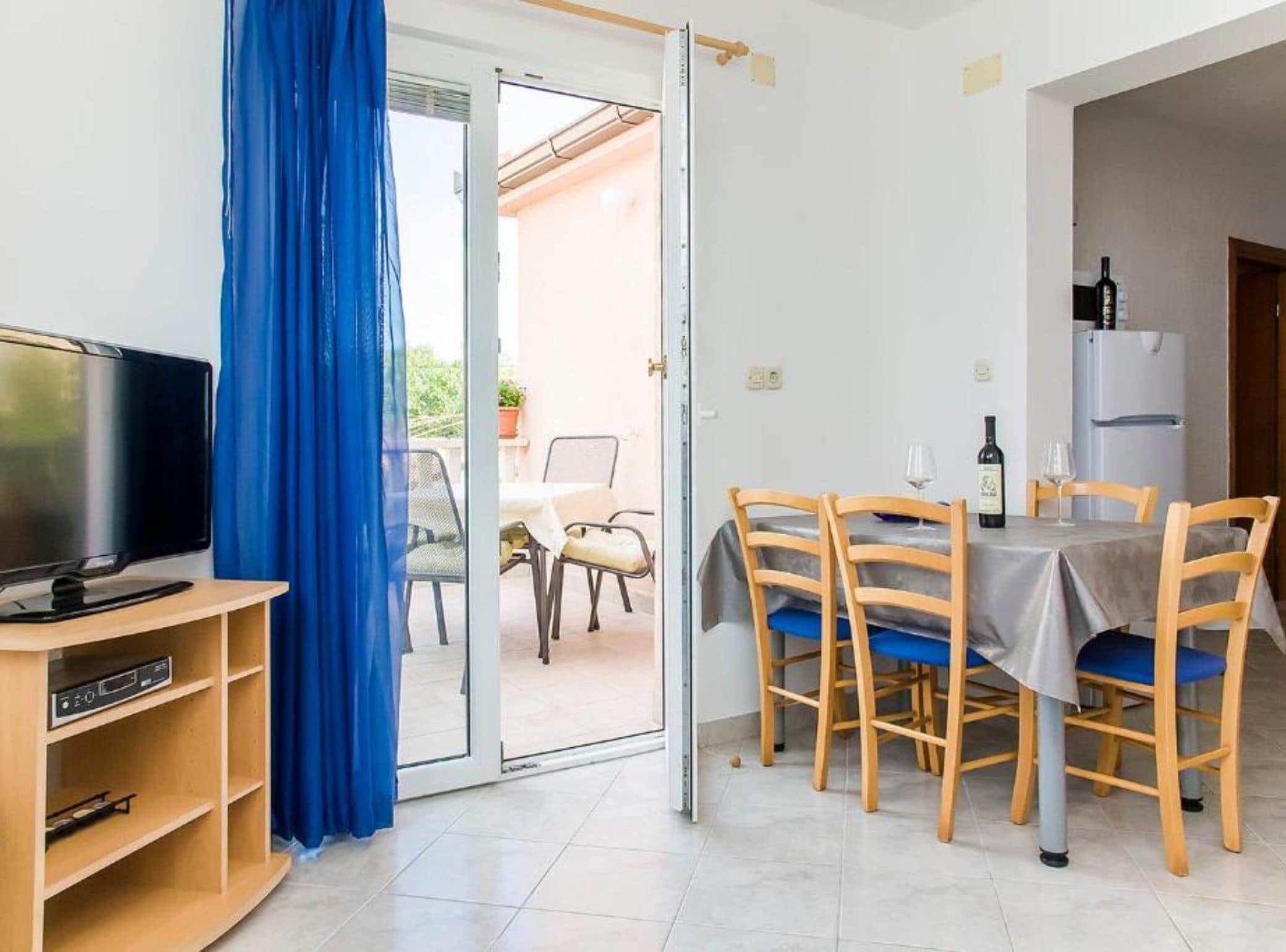 Een kamer met een televisie, een eettafel met stoelen, een koelkast en een blauw gordijn.